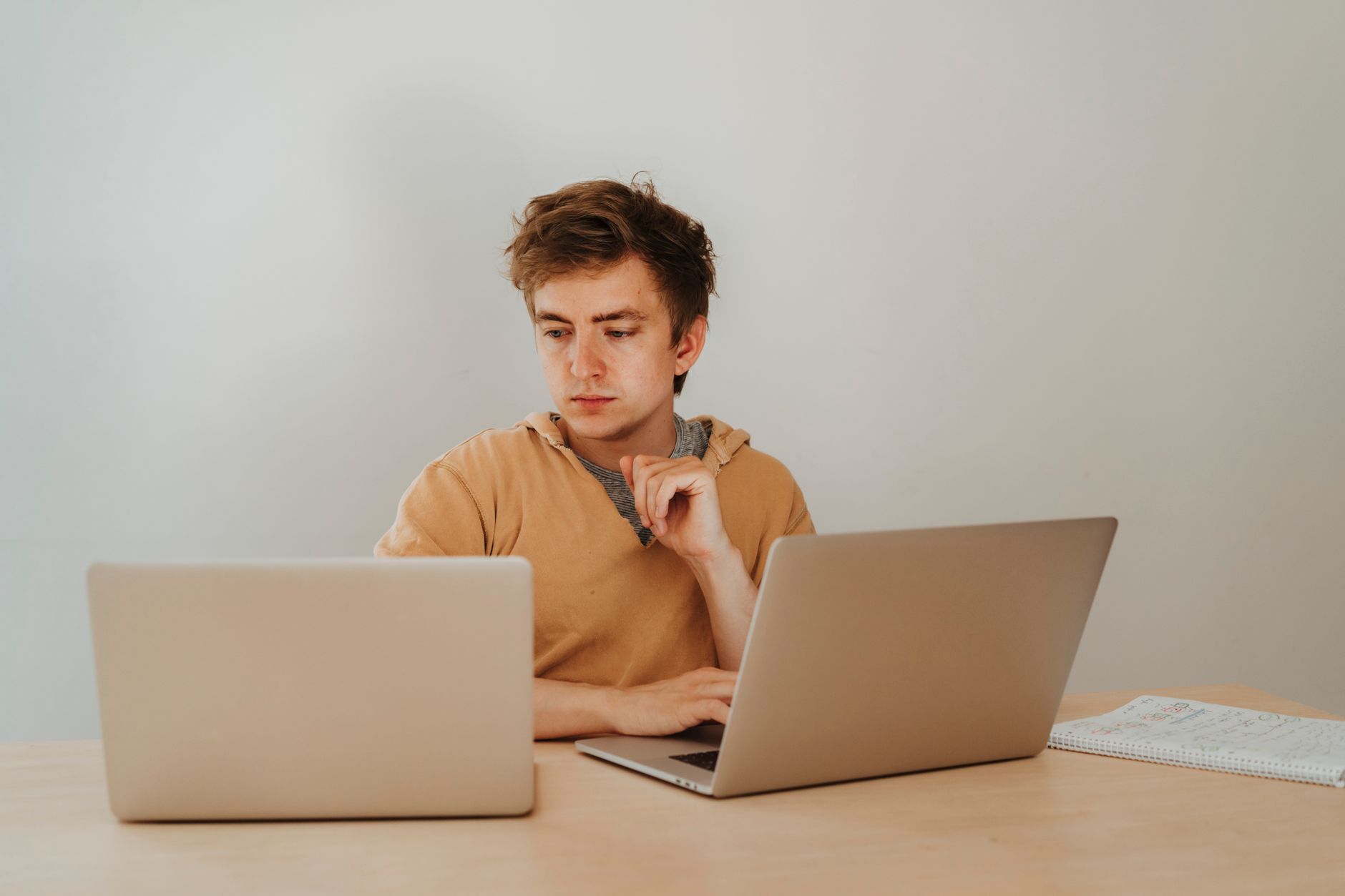 Jeune homme en sweat ocre assis à un bureau, concentré devant un ordinateur portable, avec un second portable et un cahier posés sur la table.