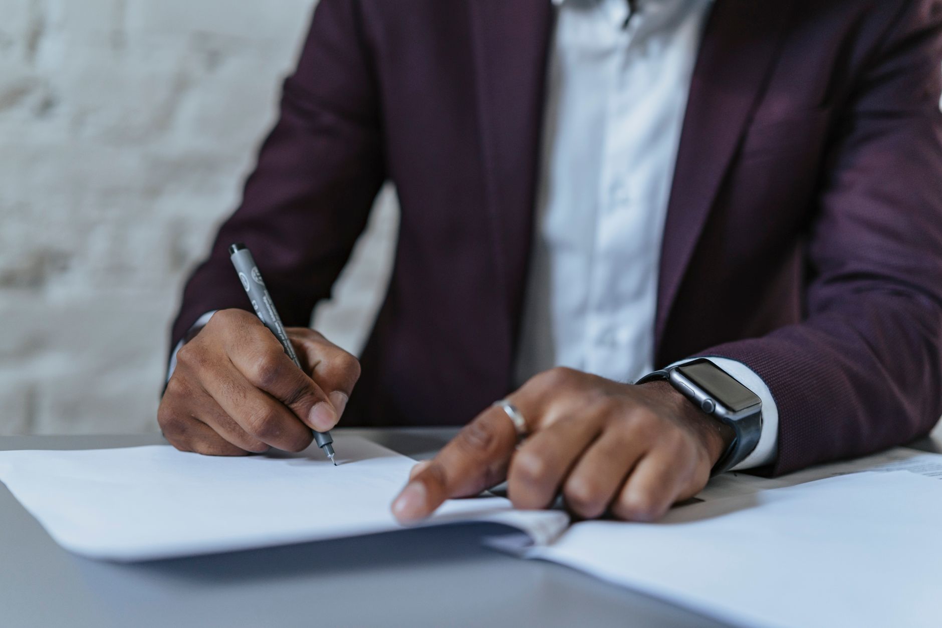 Personne en veste bordeaux signant un document à un bureau, un stylo dans une main et l’autre main posée sur la page.