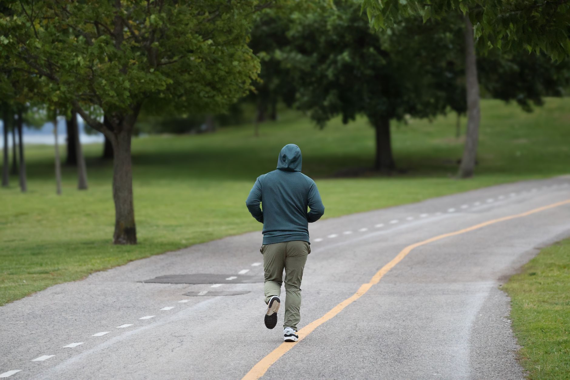 Personne vue de dos qui court sur un chemin goudronné dans un parc, en sweat à capuche vert, entre pelouse et arbres.