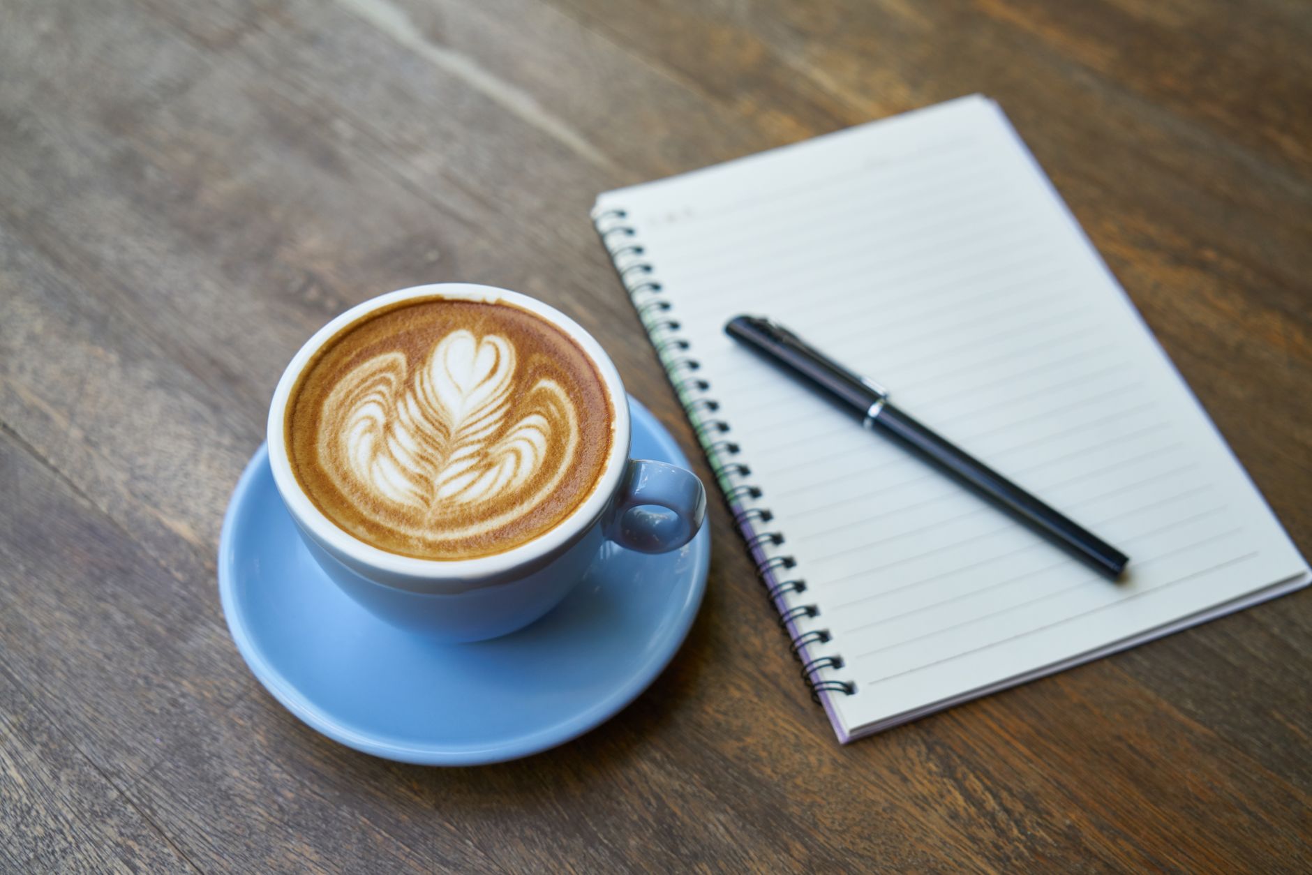 Tasse de café bleue sur soucoupe avec mousse décorée, carnet à spirale vierge et stylo noir posés sur une table en bois.