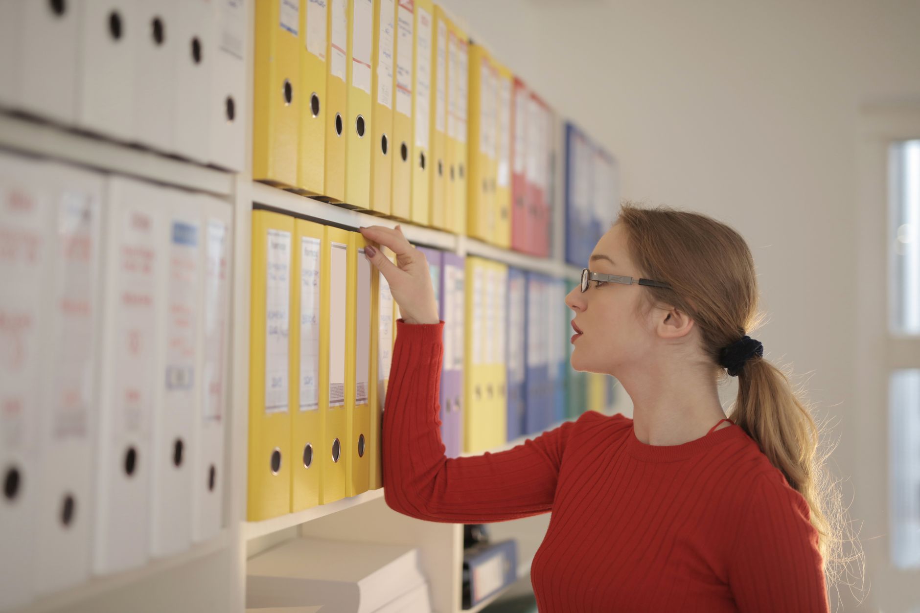 Une femme en pull rouge et lunettes attrape un classeur jaune sur une étagère remplie de classeurs de bureau.