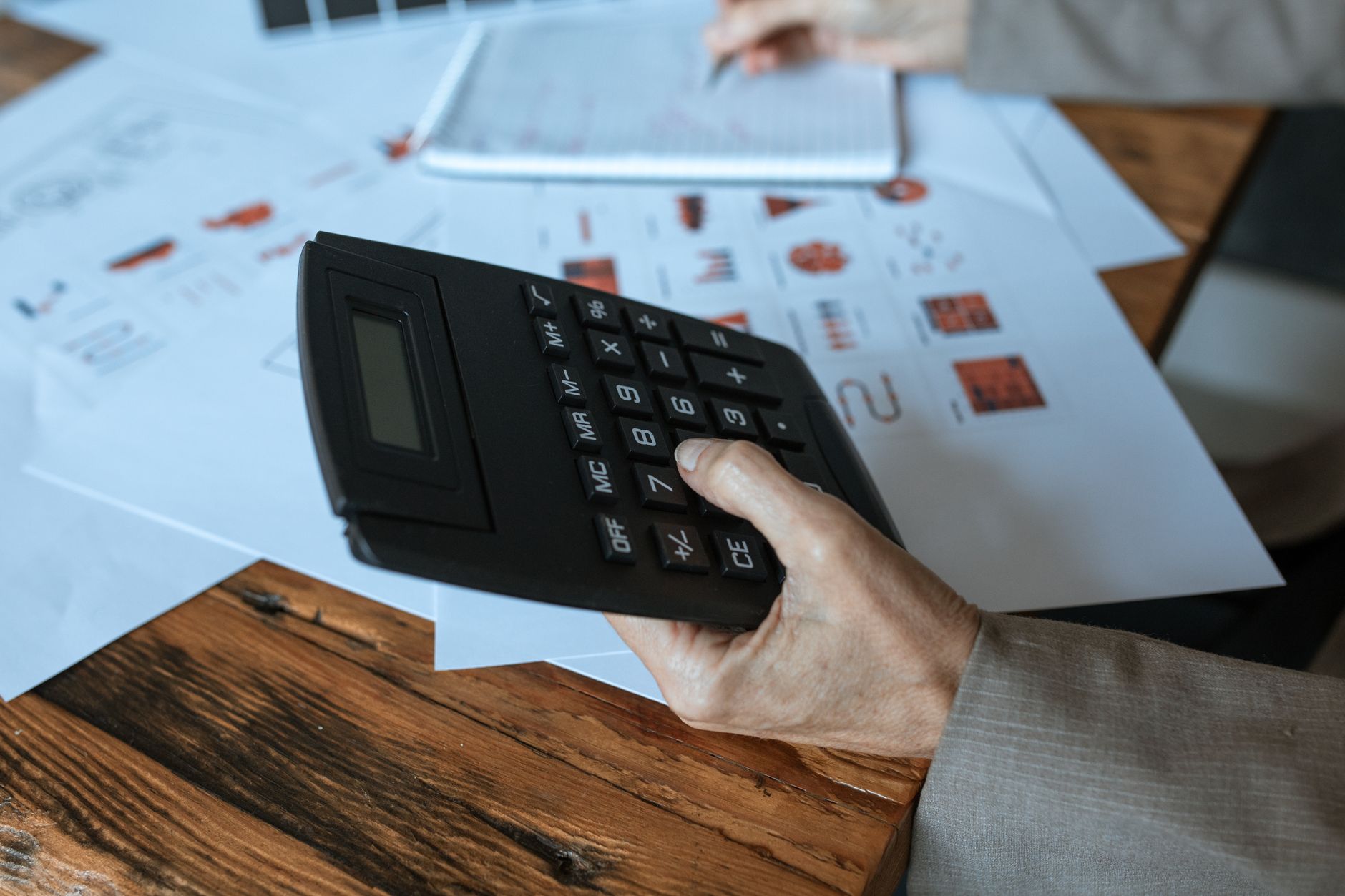 Une main tient une calculatrice noire au-dessus de documents graphiques posés sur un bureau en bois, avec un bloc-notes en arrière-plan.
